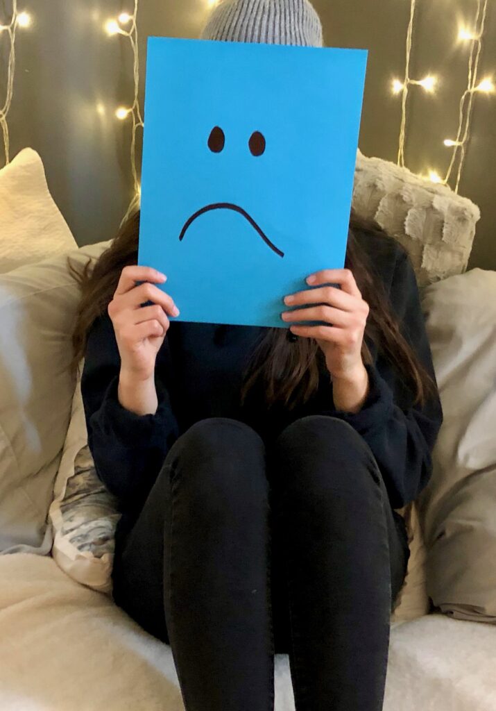 person covering their face with a blue piece of paper with a sad face on it