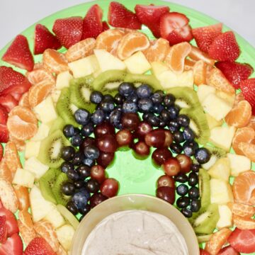 Red strawberries, Orange orange slices, Yellow pineapple, Green kiwi, Blue blueberries, and Purple grapes arranged to look like a rainbow on green platter with gold colored bowl full of dip