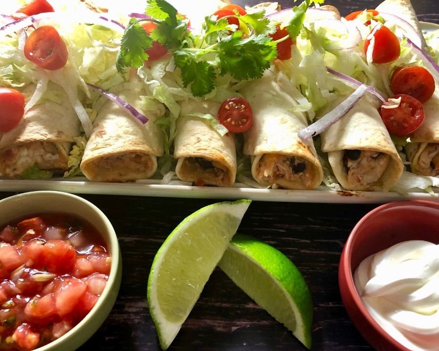 A platter full of flautas garnished with tomato, red onion, cilantro, salsa, sour cream, and lime wedges