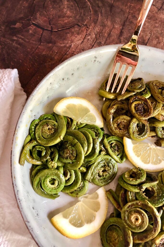 Cooked fiddleheads in a plate garnished with lemon wedges, gold fork, white napkin on side on brown table