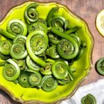 Fiddlehead ferns in a green bowl on brown table with white napkin and lemon wedges to side