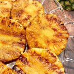Grilled Pineapple on a glass platter on a wooden table with a pineapple top in the right upper corner