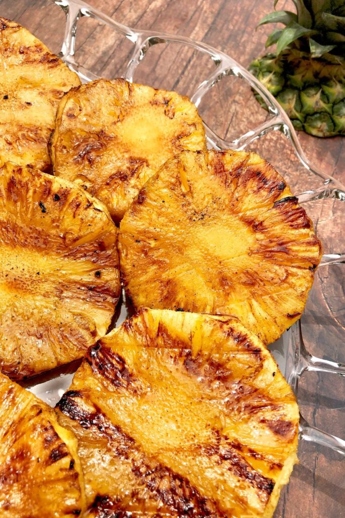 Grilled pineapple slices on glass platter on wooden table with pineapple top off to right upper corner