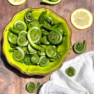 Fiddlehead ferns in a green bowl on a brown table with lemons and white napkin the sides