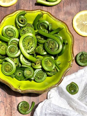 Fiddlehead ferns in a green bowl on a brown table with lemons and white napkin the sides