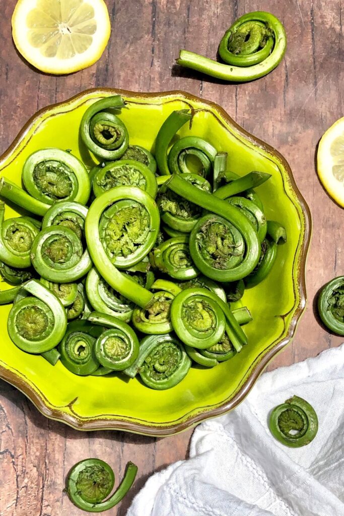 Fiddlehead ferns in a green bowl on brown table with lemon wedges and white napkin to side