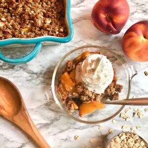 serving of peach crisp with a spoon surrounded by baking dish of peach crisp, 2 peaches, wooden spoon, and scattered oats