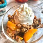 serving of peach crisp with a spoon surrounded by baking dish of peach crisp, 2 peaches, wooden spoon, and scattered oats