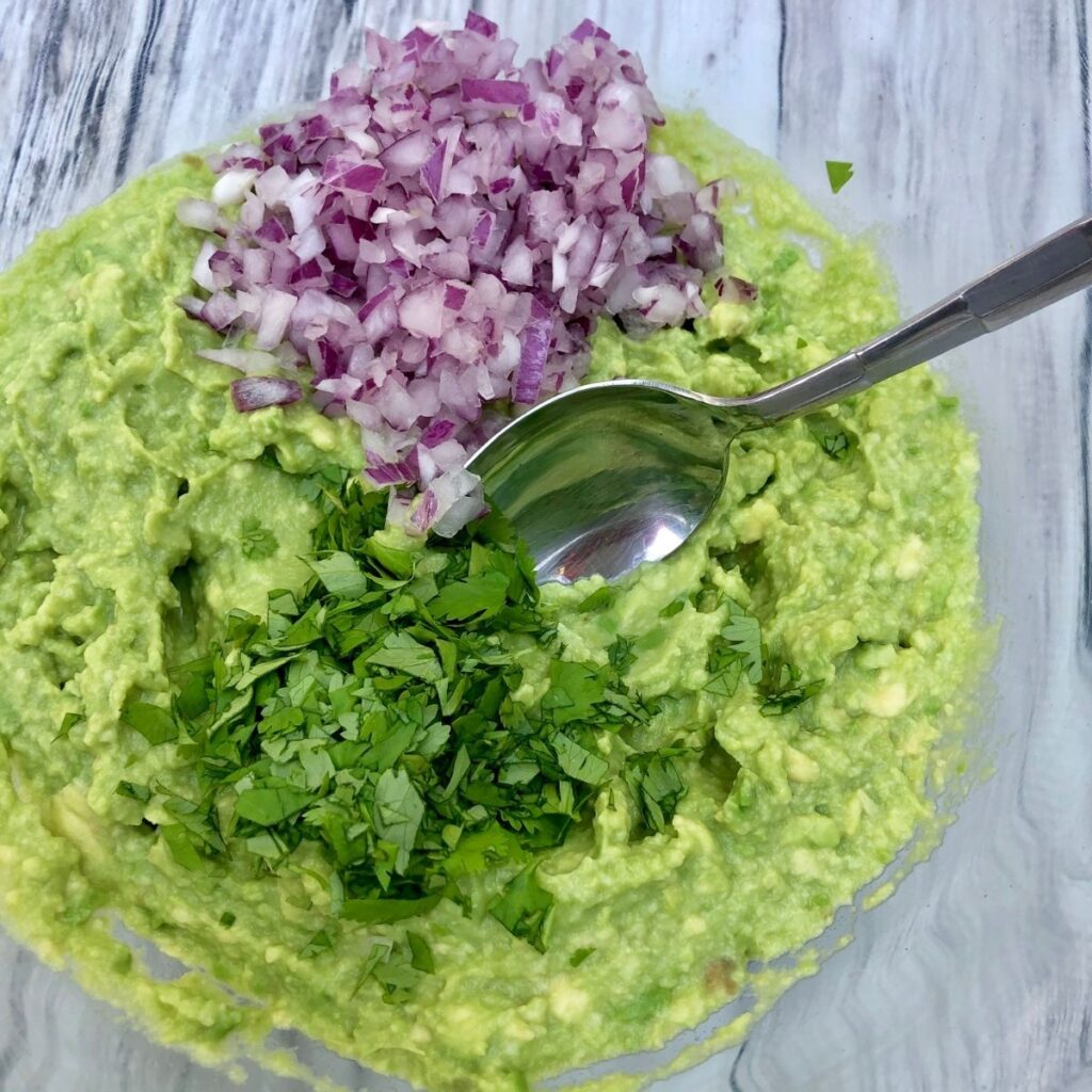 Bowl of guacamole with red onion and cilantro being stirred in