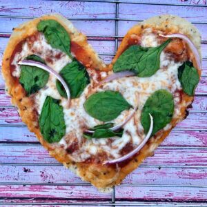 Homemade heart shaped pizza with spinach and red onion on wire rack over pink wooden background