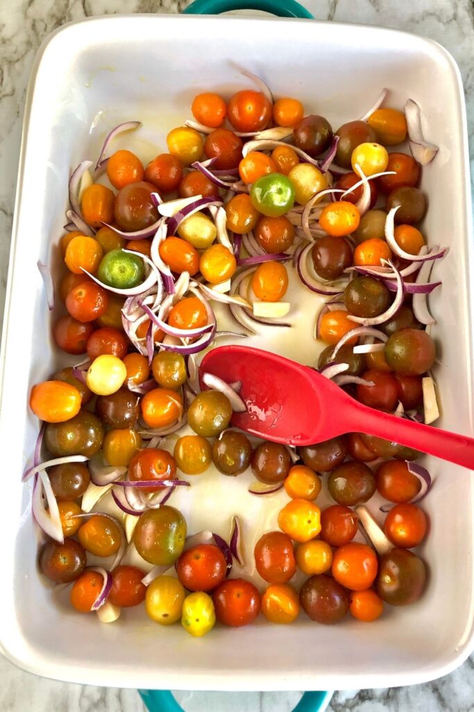 multi-colored cherry tomatoes, sliced red onion, and olive oil being mixed in a 9x13 white dish with red spoon