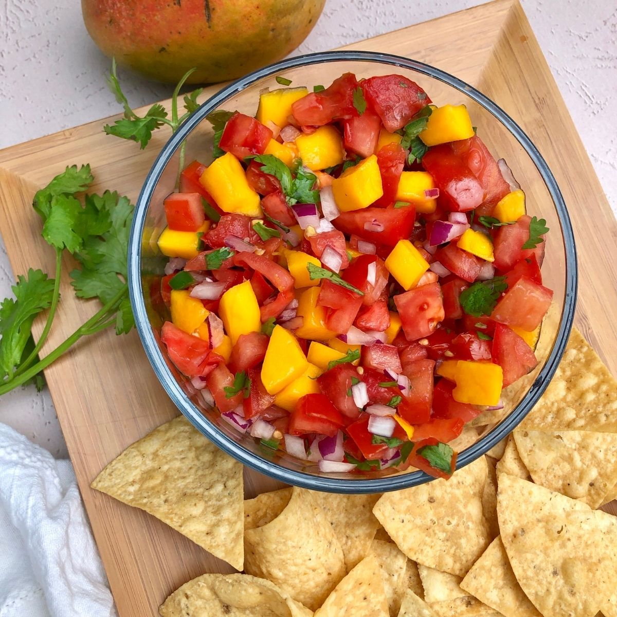 bowl of Mango Pico De Gallo on a wooden platter garnished with tortilla chips, fresh cilantro with a fresh mango off to the side