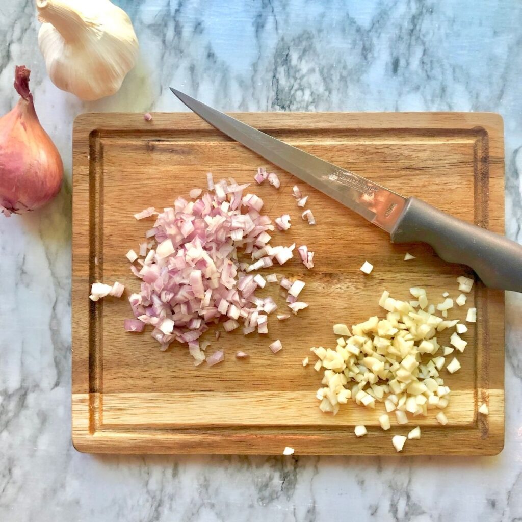 a wooden cutting board with minced shallot and garlic and a knife to the side