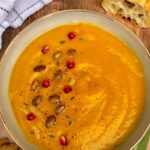 a bowl of butternut squash soup garnished with pomegranate and pumpkin seeds with crusty bread off to side