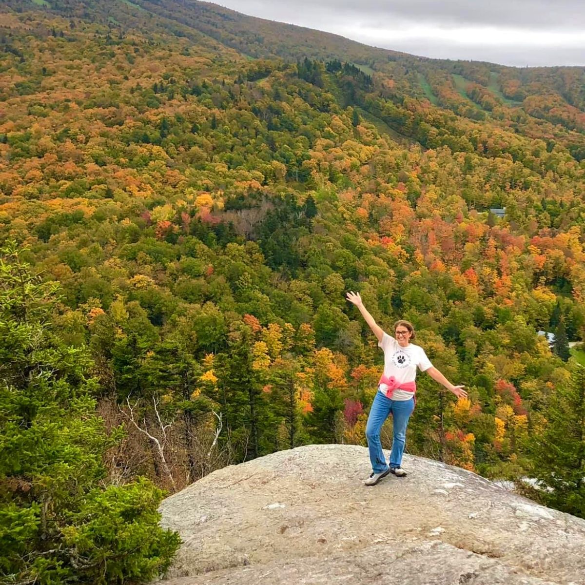 Women on a rock on a mountain with her arms stretched out, fall foliage int he background