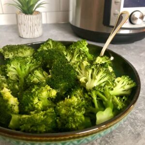 A bowl of steamed broccoli with instant pot and plant in background.