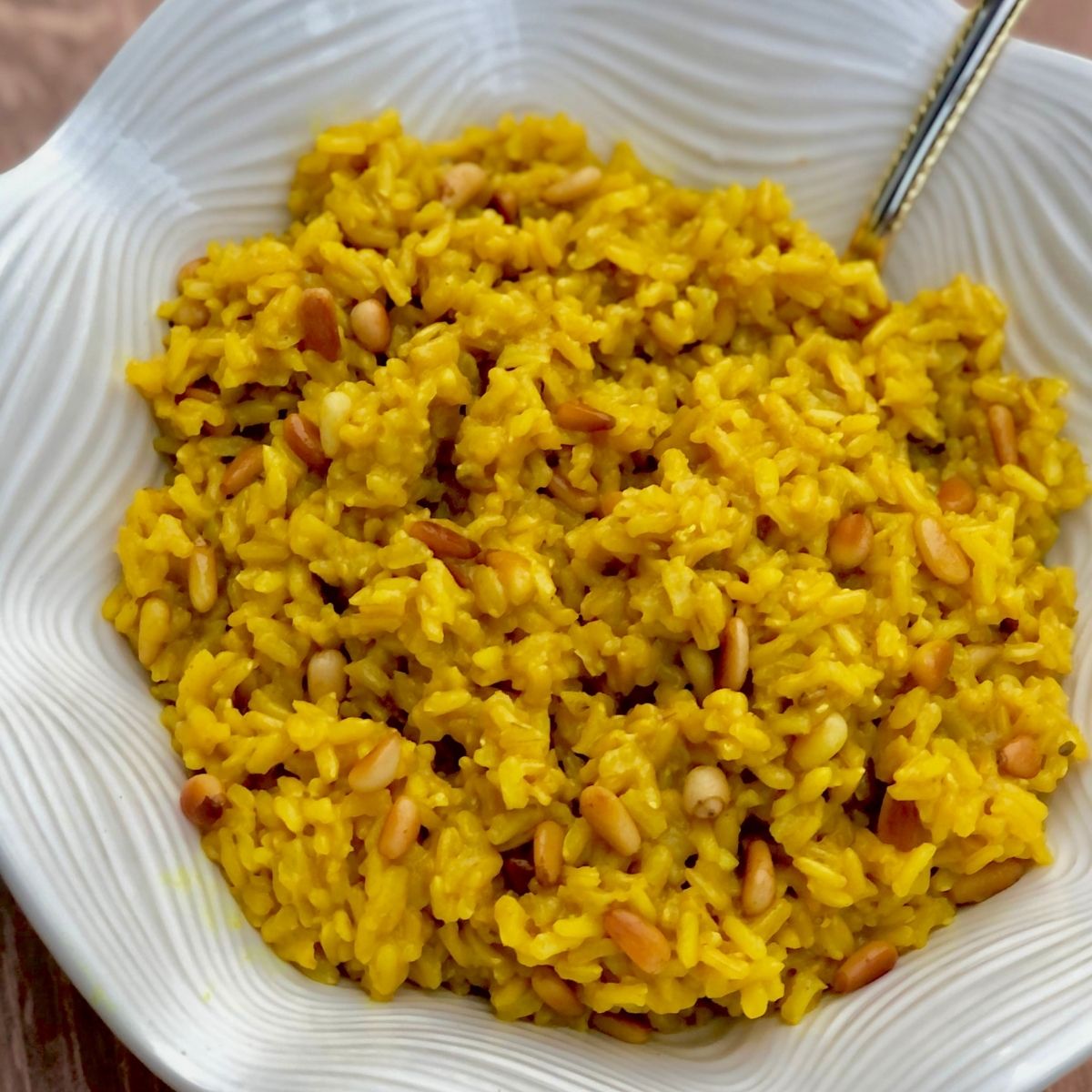 A white bowl is full of yellow colored rice with pine nuts with a sliver spoon sticking out.