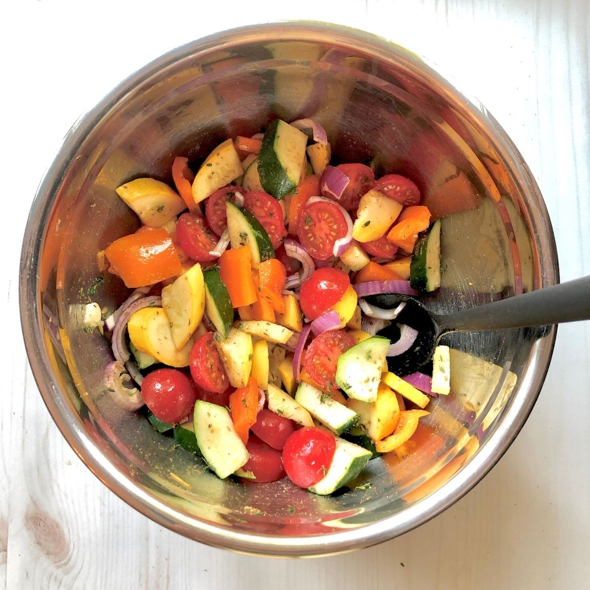 sliced tomato, zucchini, yellow squash, red onion, and orange bell peppers with olive oil and spices in a mixing bowl.
