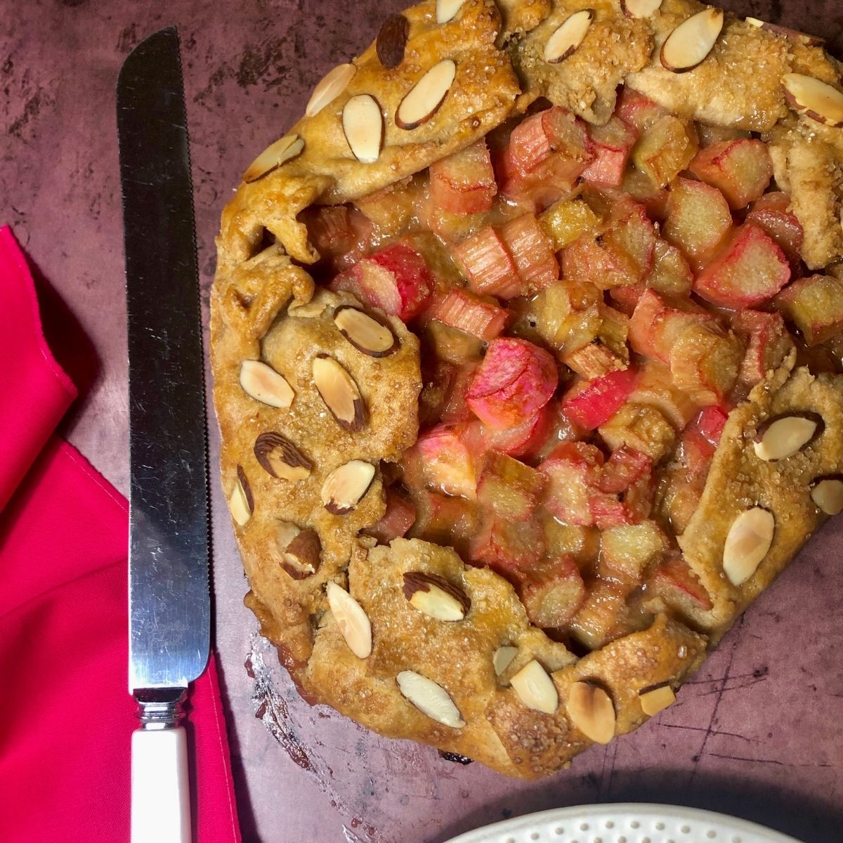 a rhubarb galette garnished with sliced almonds with a cranberry napkin and pearl handled knife to the side.
