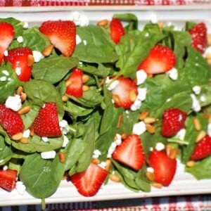 a white plate with fresh spinach, sliced strawberries, and pine nuts.