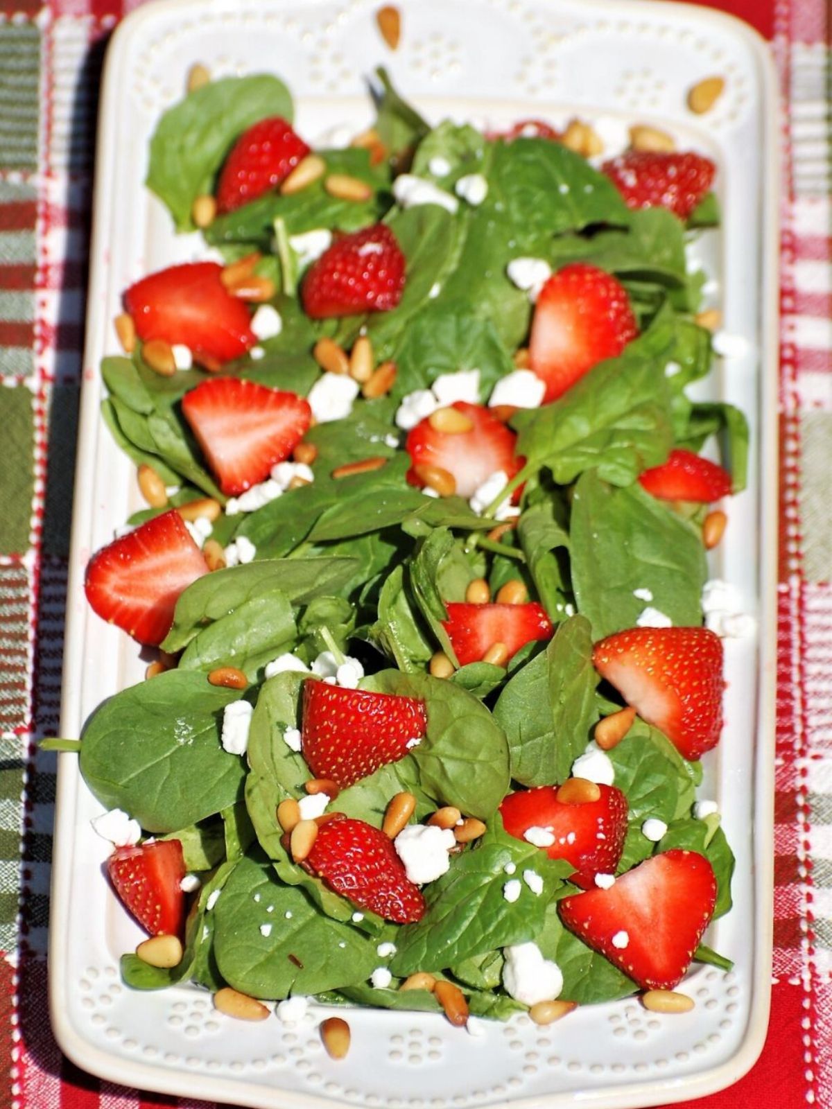 a rectangle white dish with fresh spinach, sliced strawberries, goat cheese, and pine nuts.