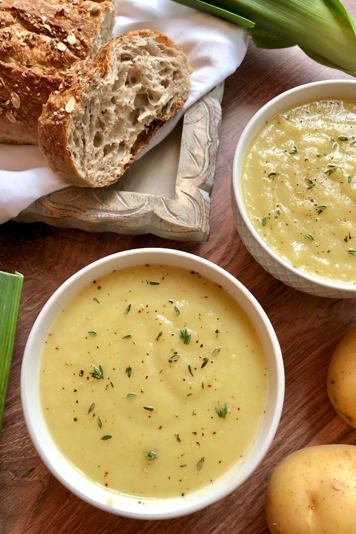 2 bowls of potato soup garnished with fresh thyme and black pepper with fresh leeks and potatoes off to side. A loaf of wheat crusty bread is on a napkin in the upper corner.