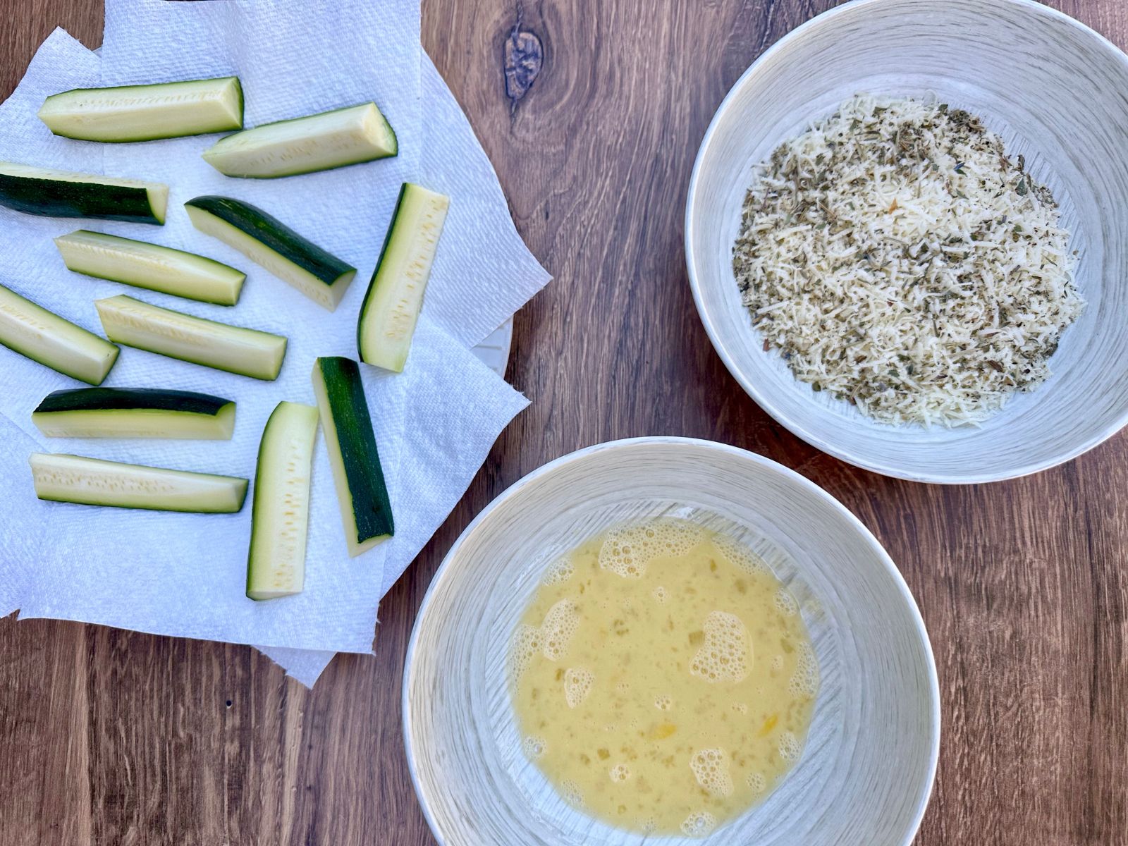 zucchini sticks on paper towels, a beaten egg in a bowl, and cheese + spices in another bowl.