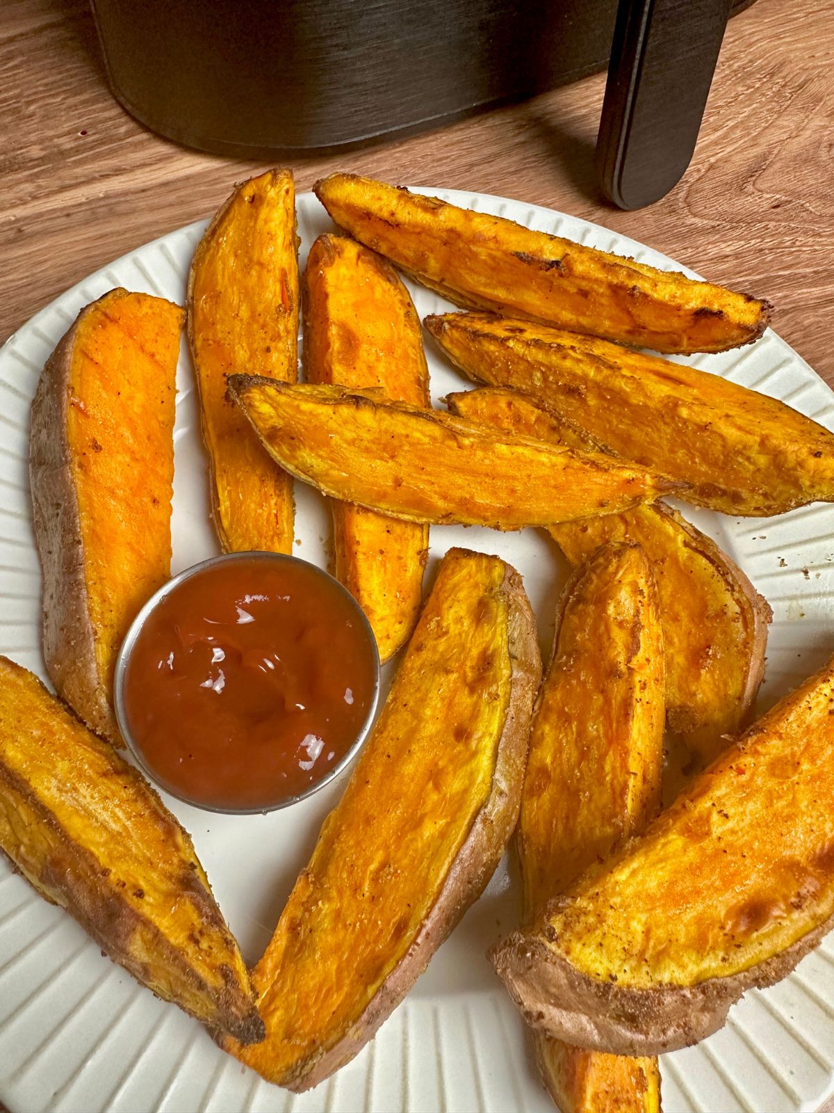 Sweet potato wedges on a white plate with condiment cup of ketchup with air fryer based in background.